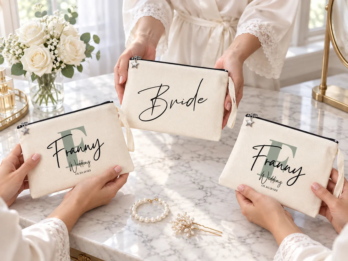 Mains de demoiselles d'honneur en peignoirs de soie sur une coiffeuse en marbre, avec une pochette « Bride » et deux pochettes « Franny Wedding », des roses blanches et un bracelet de perles