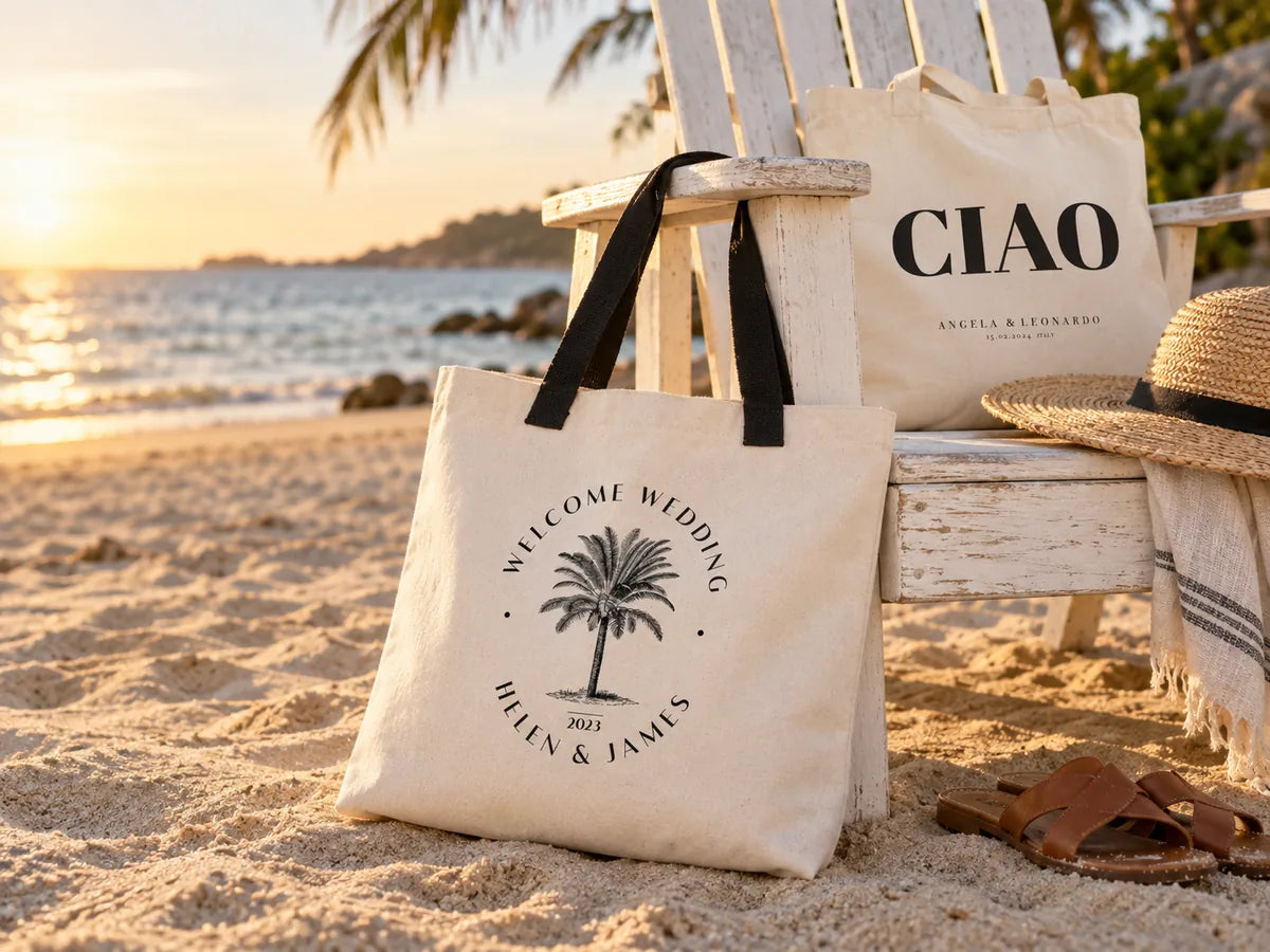 Grand sac en toile « Welcome Wedding Helen & James » à emblème palmier et anses noires, appuyé contre une chaise Adirondack blanche sur la plage à l'heure dorée, avec un sac CIAO, un chapeau de paille et des sandales en cuir